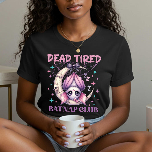 Dead tired and fully over it? Same. Join the Batnap Club with this pastel goth bat Halloween shirt, featuring a sleepy little winged gremlin who’s clearly clocked out. Wrapped in a pink bow and hanging like tomorrow’s problems, this adorable burnout bat is all of us in October (and every Monday). Perfect for spooky naps, haunted errands, or just surviving on caffeine and vibes. 